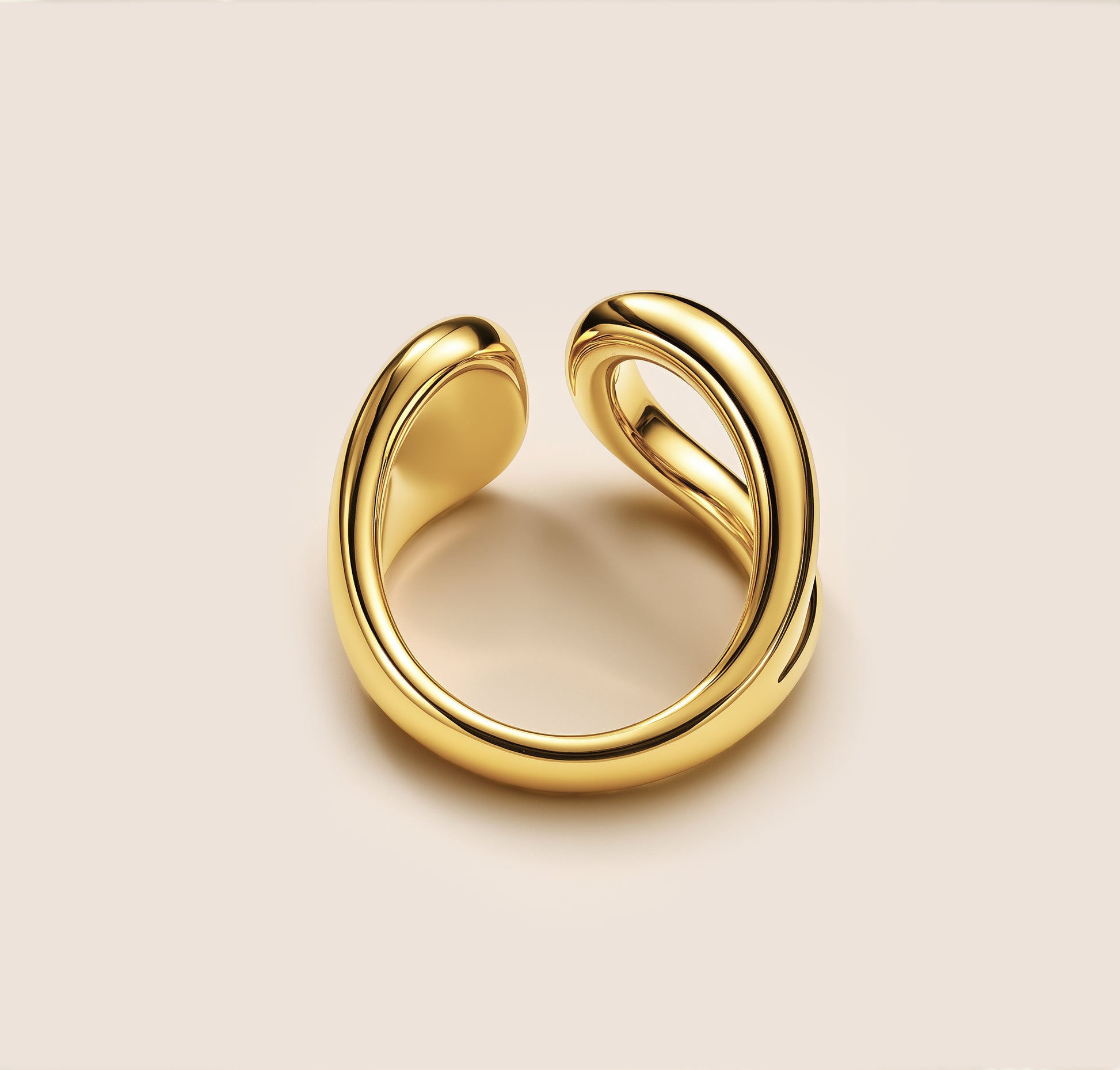 The maia ring, a gold ring with an open, sculptural design featuring two smoothly curved, interwoven bands that create a fluid, modern look. The high-polish finish reflects light, set against a soft beige background.